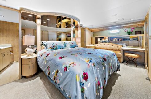 Yacht bedroom with colorful bedding, sofa, desk with chair, storage cabinets, and view into bathroom