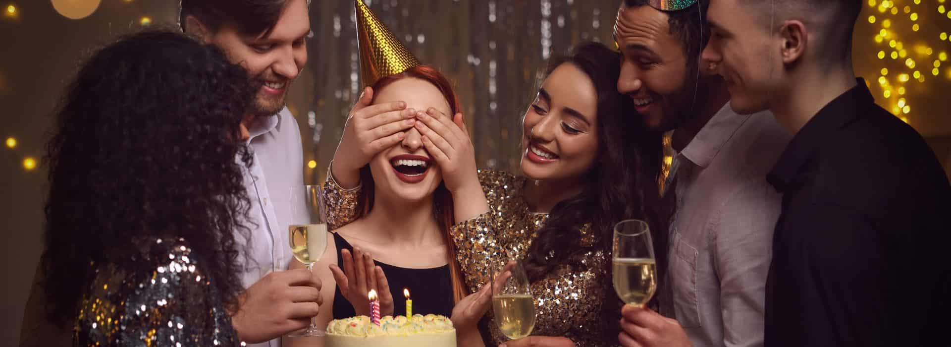 People dressed up for a birthday party surrounding the birthday girl in front of a cake with her eyes covered