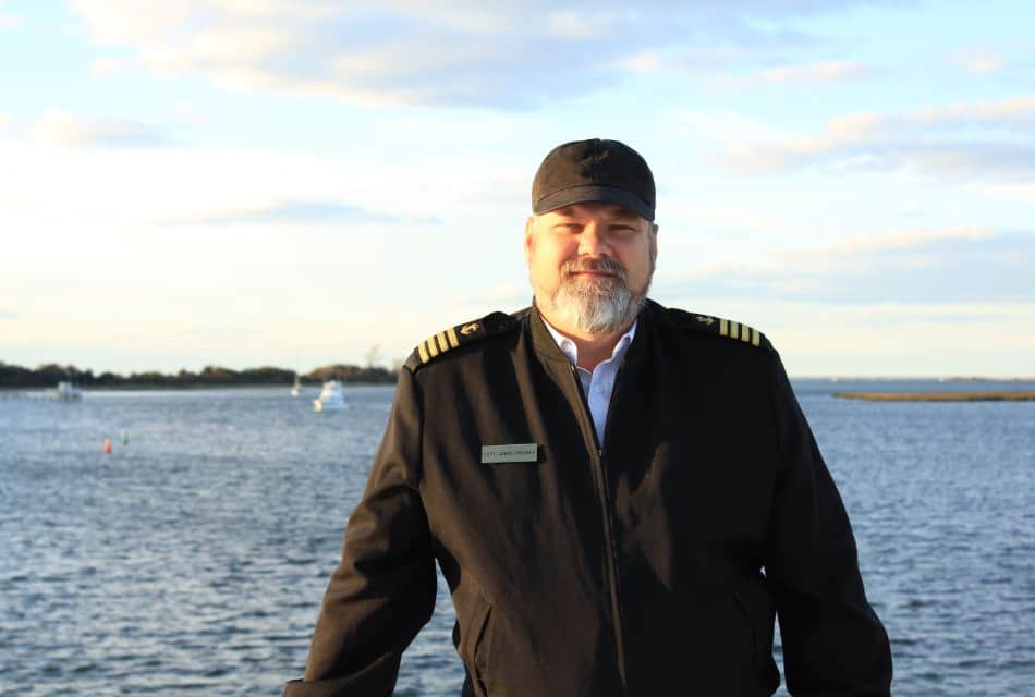 Man with black boat captain's hat and jacket with view of water behind him