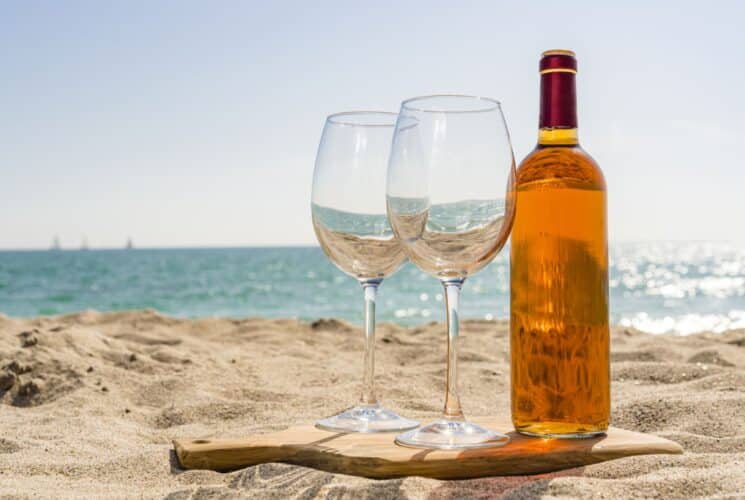 Close up view of two wine glasses and a bottle of wine on a wooden cutting board on the beach sand with the ocean in the background